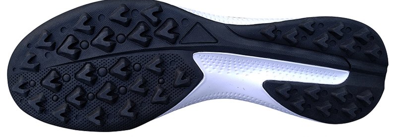 A black turf futsal sole seen from below: rows of short chevron-shaped rubber studs, with a white cushioning layer showing through the middle of the sole.
